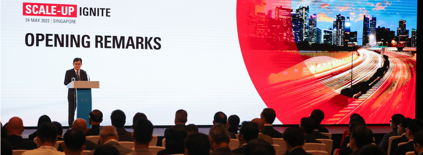 "Scale-Up Ignite" event: Speaker at lectern before audience and cityscape backdrop.